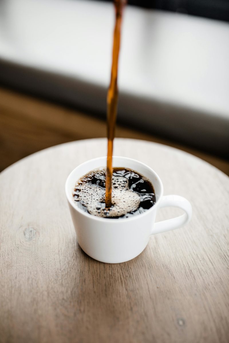 Steaming black coffee being poured into a white ceramic mug on a wooden table.