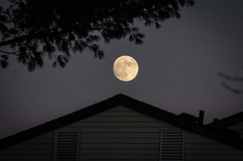 A full moon illuminating the night sky above a silhouetted house with trees.