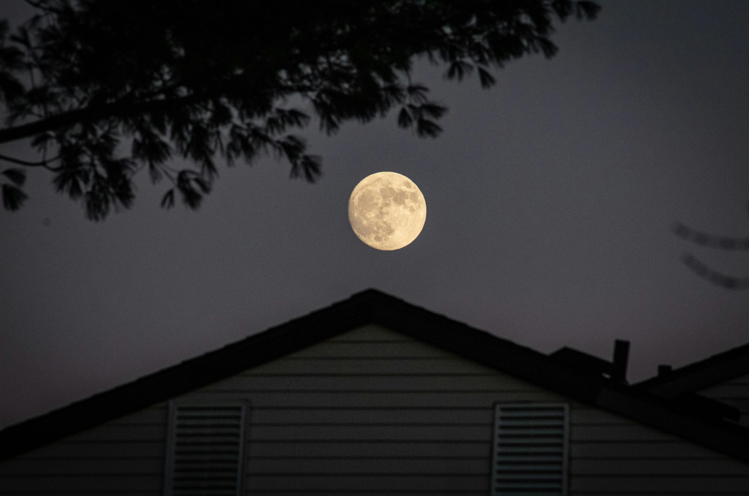 A full moon illuminating the night sky above a silhouetted house with trees.